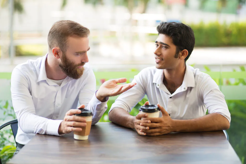 two men are discussing something at a table with a cup of coffee each